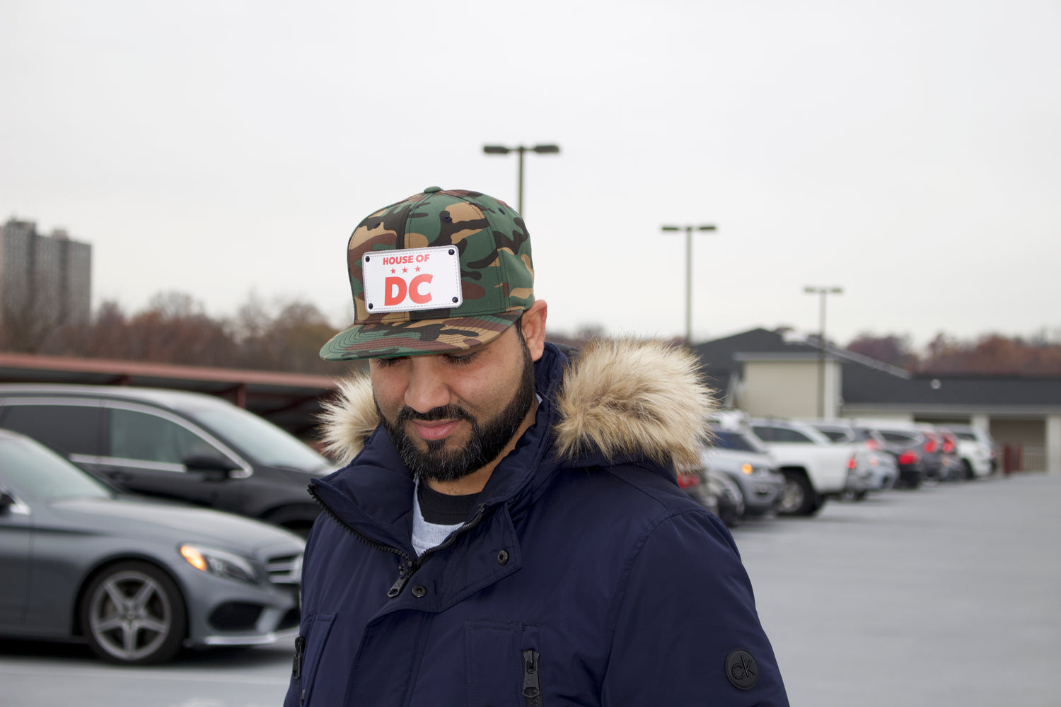 Man wearing a camouflage HOUSE OF DC hat with a sticker and a fur-lined coat in a parking lot.