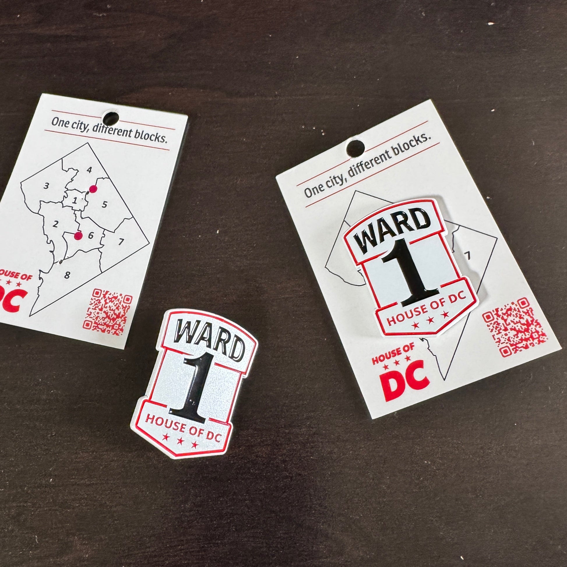 Ward 1 House of DC enamel pin shown with its backing card reading 'One city, different blocks,' featuring a map of DC's eight wards and a QR code, alongside the red, black, and white shield-shaped pin.