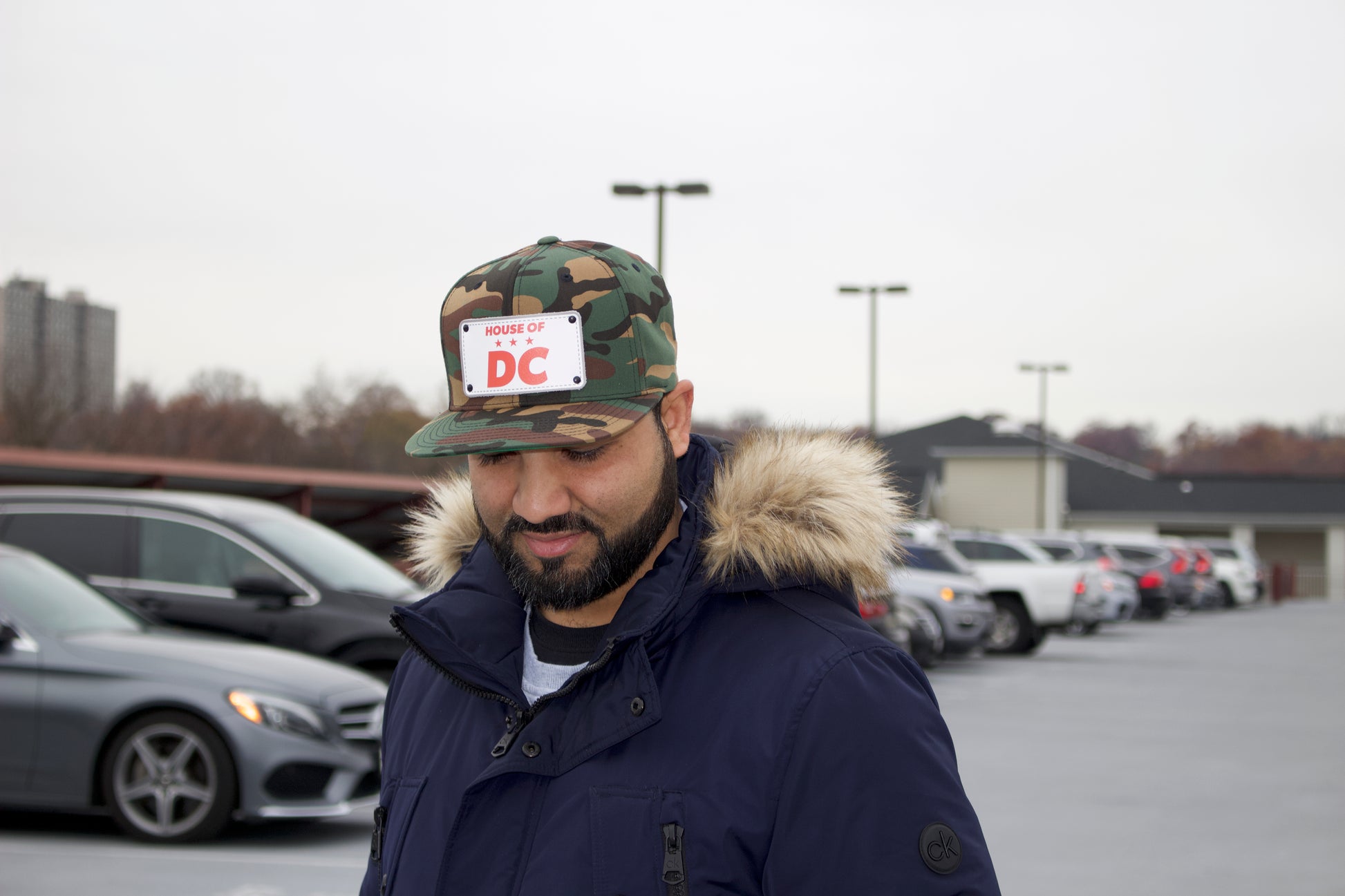 Man wearing a camouflage HOUSE OF DC hat with a sticker and a fur-lined coat in a parking lot.