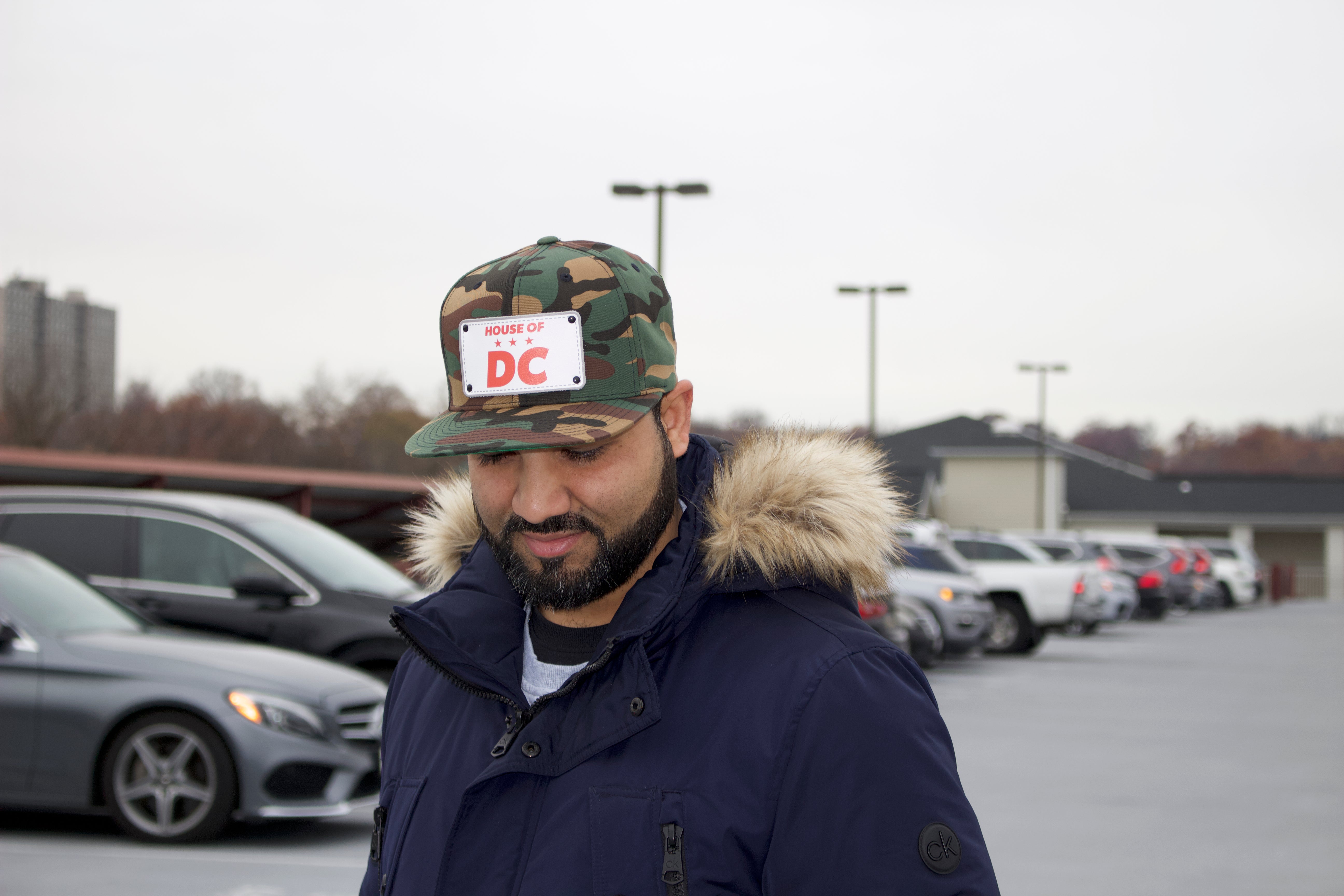Man wearing a camouflage HOUSE OF DC hat with a sticker and a fur-lined coat in a parking lot.