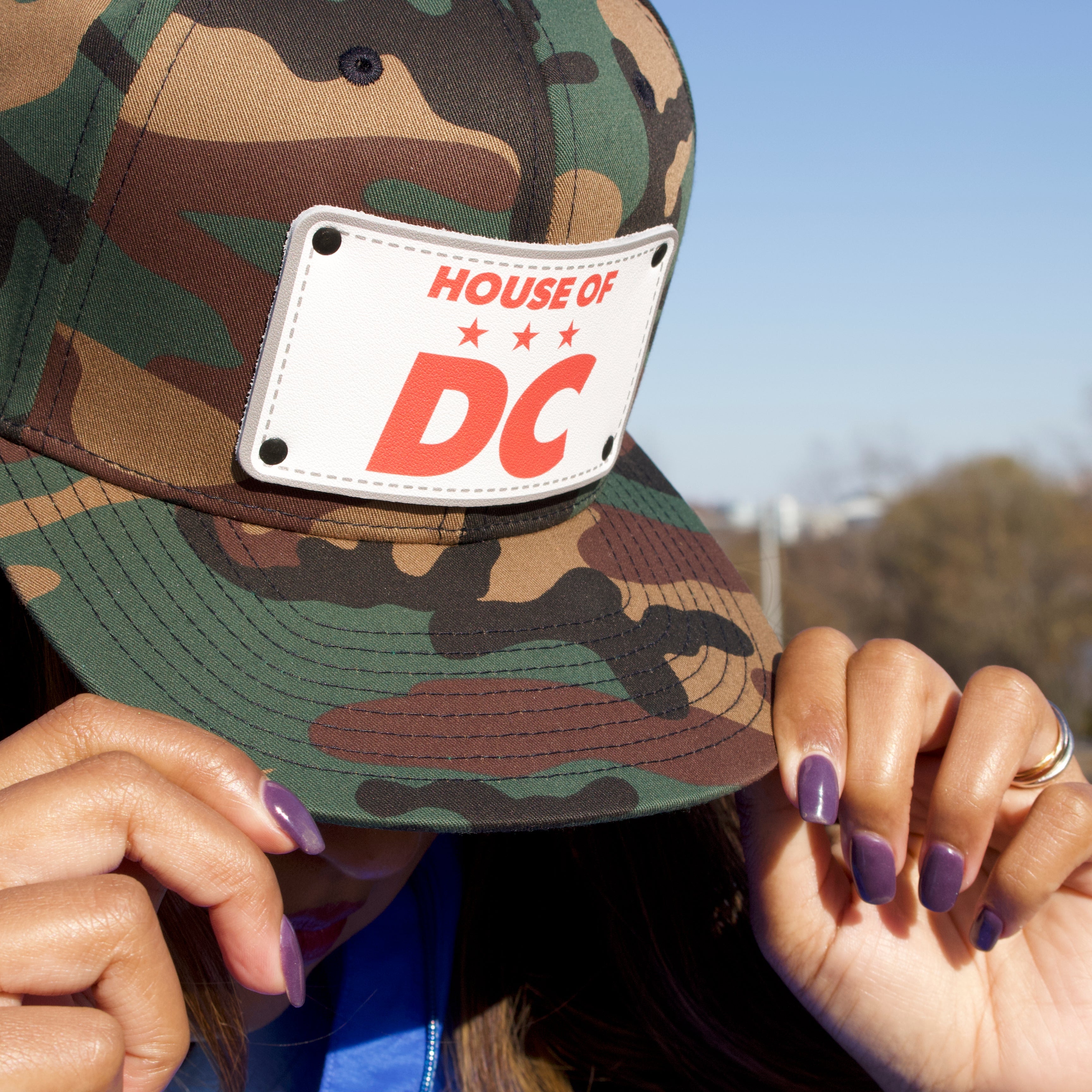 Camo snapback hat in camo that says HOUSE OF DC on female with purple fingernails. 