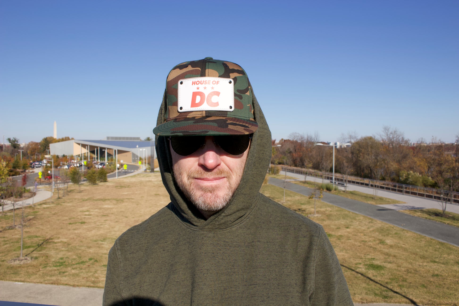 HOUSE OF DC KB, man wearing camo hat with green hoodie outdoors with Washington Monument in the background. 