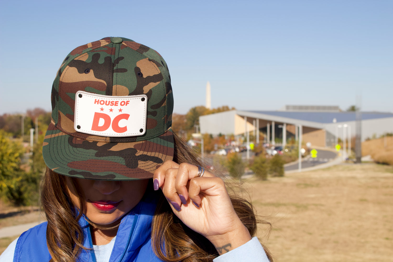 Woman wearing a HOUSE OF DC camo snapback hat with purple fingernails and red lipstick and Washington city in background. 