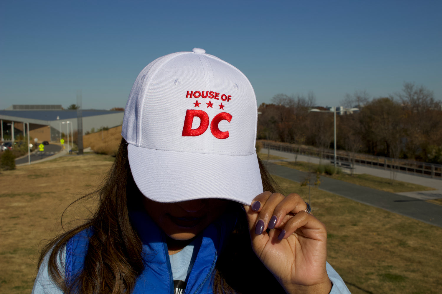 Woman wearing a HOUSE OF DC white baseball cap with purple fingernails and red lipstick and Washington in background. 